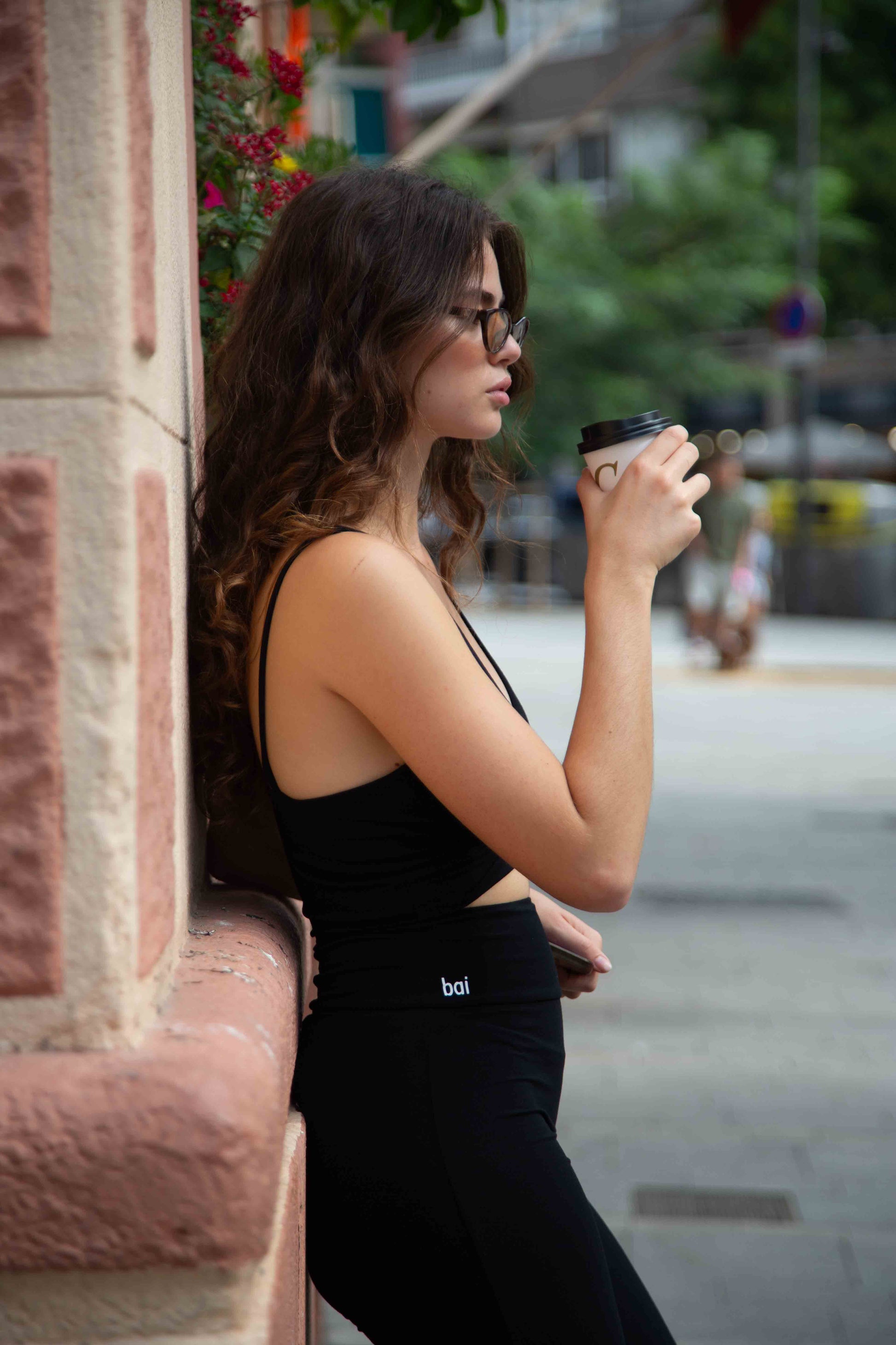 Woman in black outfit drinking from a coffee cup on a city street.