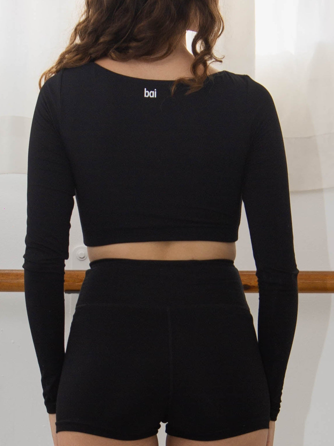 Person wearing a black outfit in a dance studio with white curtains and a barre in a longsleeve crop top.