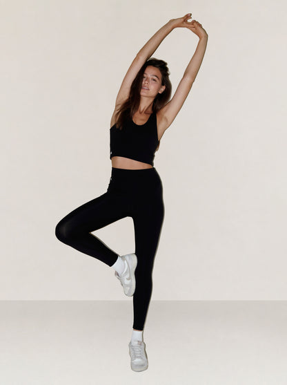 A woman in a black natural fibre sports bra and black leggings poses in a yoga tree pose while wearing white sneakers against a plain, light beige studio background.
