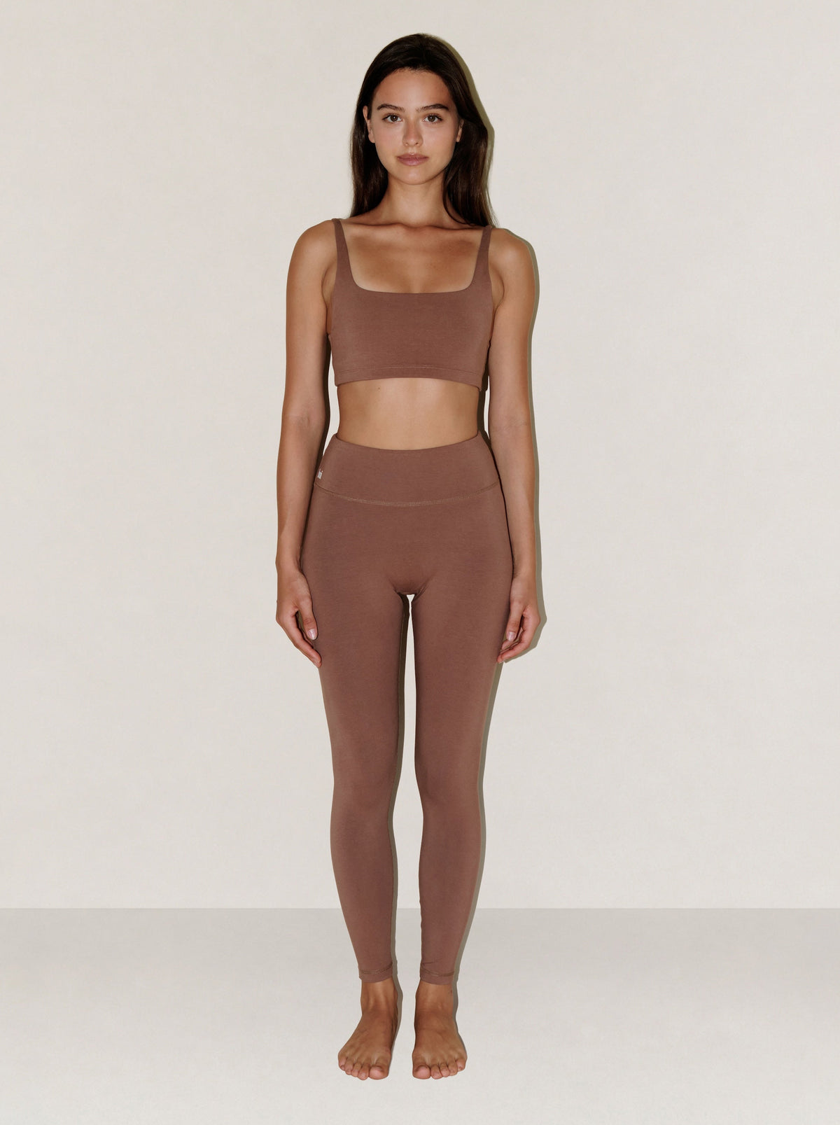 A woman stands facing forward in a matching two-piece activewear set consisting of a square-neck sports bra and high-waisted leggings in a solid taupe-brown color, set against a plain light gray studio background.