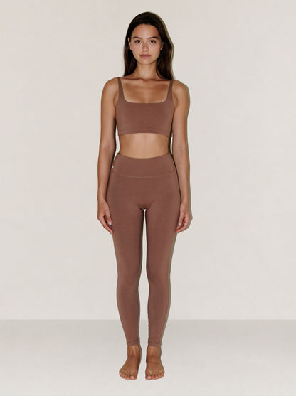 A woman stands facing forward in a matching two-piece activewear set consisting of a square-neck sports bra and high-waisted leggings in a solid taupe-brown color, set against a plain light gray studio background.