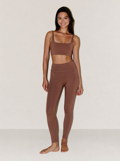 A woman modeling a matching natural fibre two-piece activewear set in a solid taupe-brown color, featuring a scoop-neck sports bra and high-waisted leggings, against a plain off-white studio background.