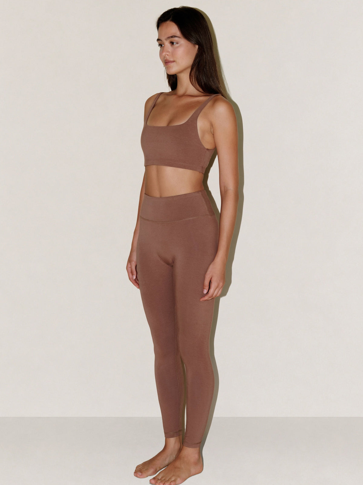 A woman stands in profile wearing a matching natural fibre two-piece activewear set in a muted taupe-brown color, consisting of a scoop-neck crop top and high-waisted leggings, against a plain off-white studio background.