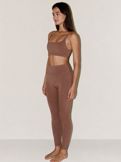 A woman stands in profile wearing a matching natural fibre two-piece activewear set in a muted taupe-brown color, consisting of a scoop-neck crop top and high-waisted leggings, against a plain off-white studio background.