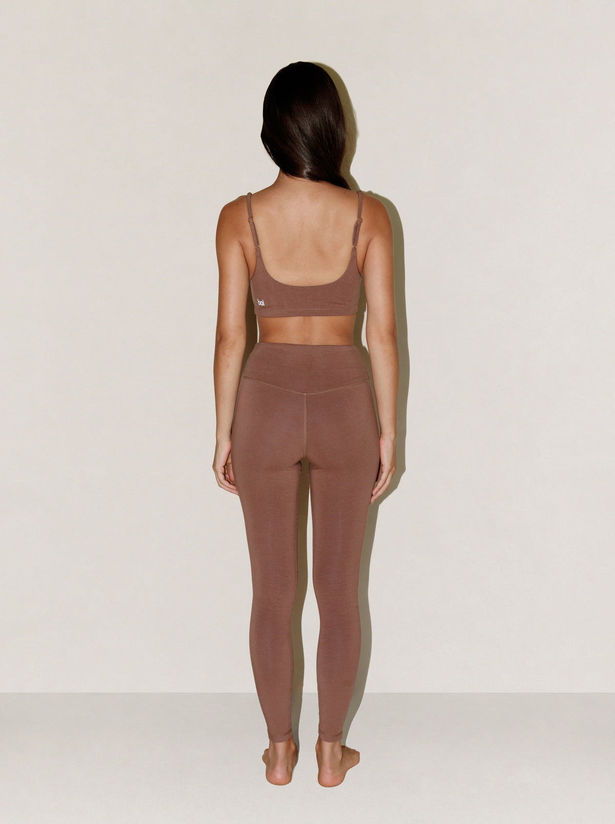 A woman stands with her back to the camera wearing a matching natural fibre two-piece activewear set in a muted taupe-brown color, consisting of a sports bra and high-waisted leggings, against a plain off-white background.