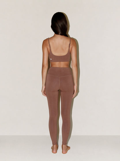 A woman stands with her back to the camera wearing a matching natural fibre two-piece activewear set in a muted taupe-brown color, consisting of a sports bra and high-waisted leggings, against a plain off-white background.