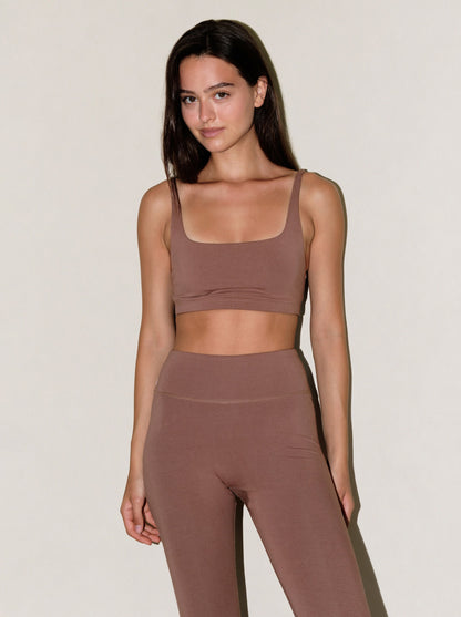 A woman modeling a matching two-piece activewear set in a dusty mauve-brown color, featuring a scoop-neck sports bra and high-waisted leggings, against a plain off-white studio background.