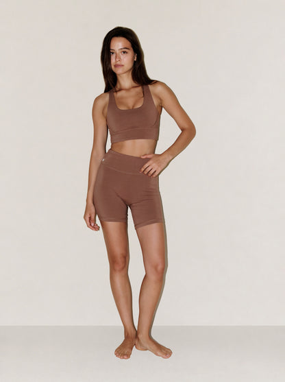 A woman modeling a matching brown natural fibre activewear set consisting of a scoop-neck sports bra and high-waisted biker shorts, standing against a plain light beige studio background. #color_brown