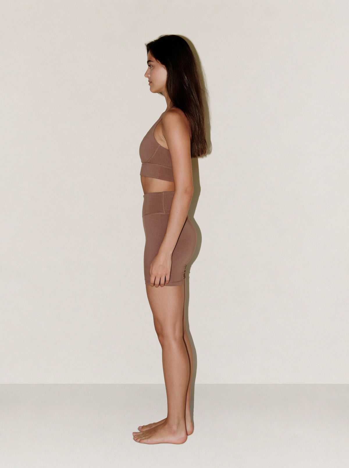 A woman stands in profile wearing a matching natural fibre mocha mousse activewear set consisting of a sports bra and high-waisted shorts, against a plain off-white studio background. #color_brown