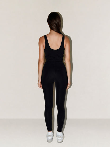 A woman viewed from behind wearing a black natural fibre sleeveless tank top and black leggings, standing against a plain off-white studio wall and floor with white sneakers.