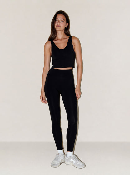 A woman modeling a black natural fibre athletic set consisting of a cropped tank top and high-waisted leggings, paired with white sneakers, against a plain off-white studio background.