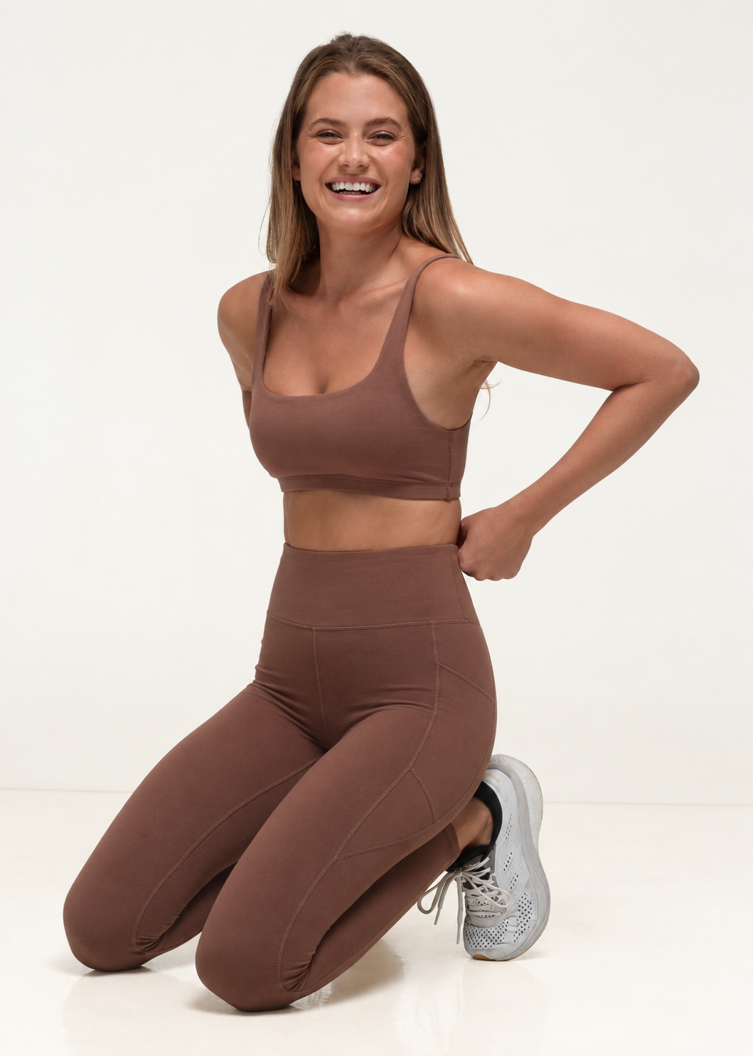Model in natural fibre activewear in mocha mousse squatting and laughing.
