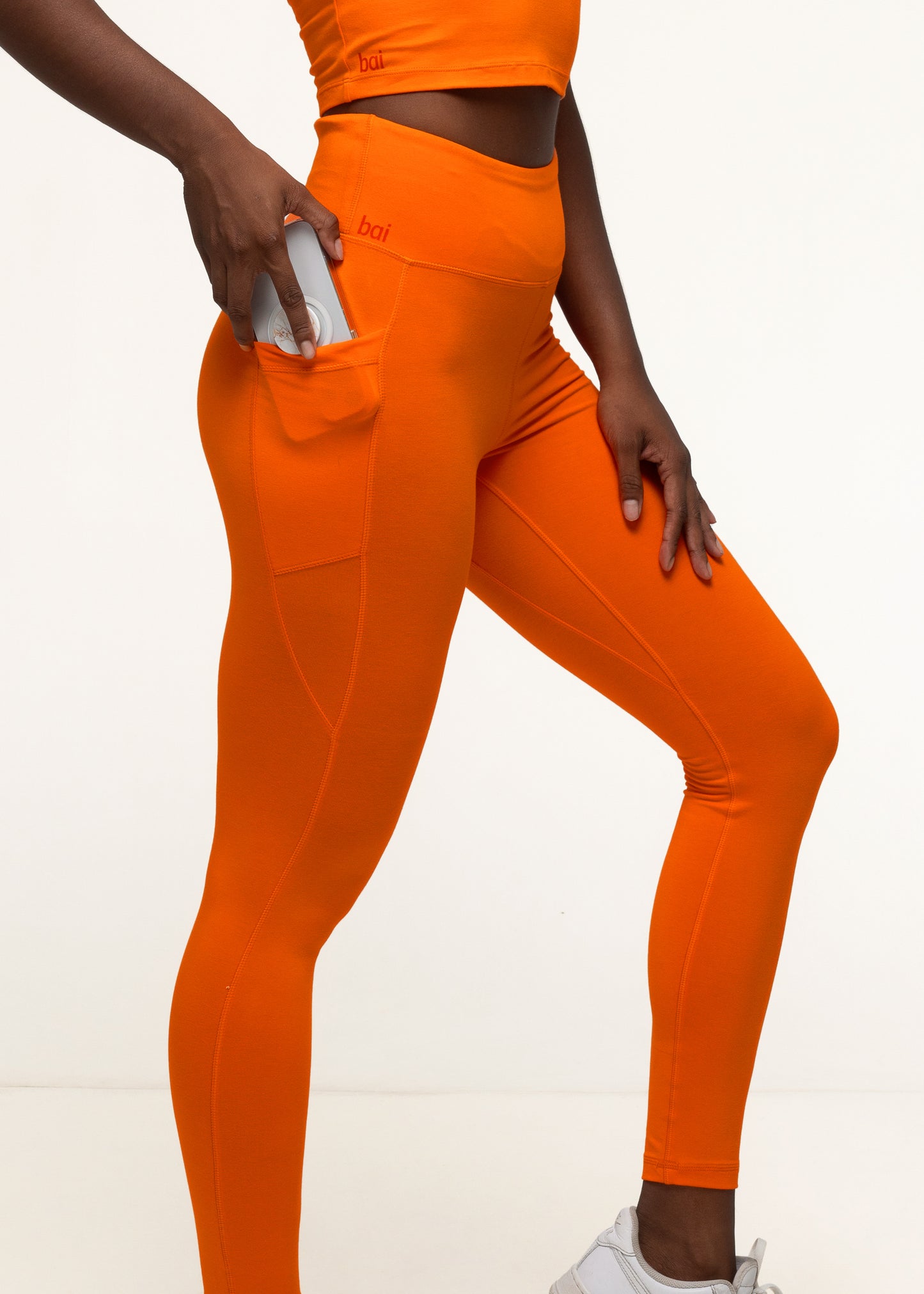 Woman securely storing a smartphone in the pocket of her bai active high-rise orange bamboo leggings, perfect for hands-free outdoor workouts. #color_orange