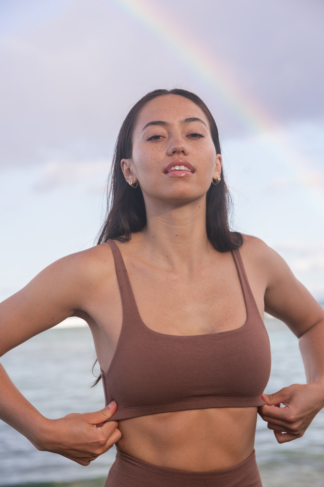A woman wearing a brown sports bra and matching bottoms stands outdoors with her hands on her hips, pulling the sports bra up slightly to show the fit. A soft rainbow is visible in the sky behind her, and water is in the background.